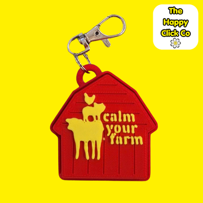 A red barn shaped  fidget clicker keychain with  yellow farm animals and calm your farm text in the center and a silver clasp at the top, shown on a bright yellow background. A 'The Happy Click Co' logo is visible in the top right corner.