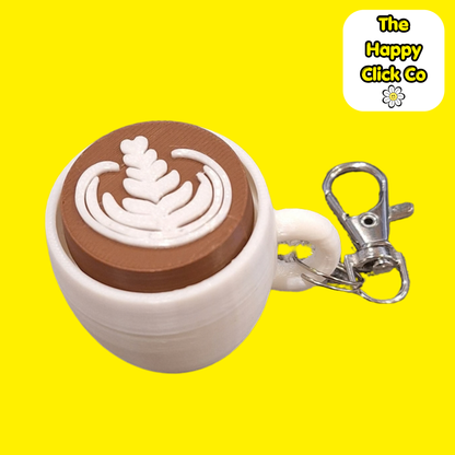 A white coffee cup shaped fidget clicker keychain with a brown centre and cappuccino milk pattern and a silver clasp at the top, shown on a bright yellow background. A 'The Happy Click Co' logo is visible in the top right corner.