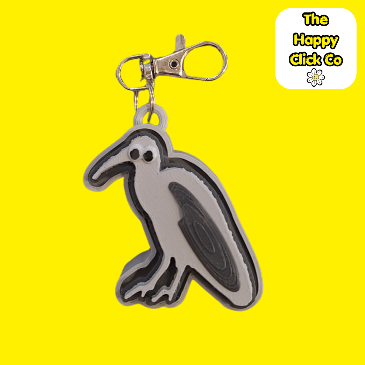 A South African inspired grey and  black Hadeda bird shaped fidget clicker keychain with a silver metal clasp, featuring cartoon eyes on a bright yellow background. A logo for 'The Happy Click Co' is visible in the top right corner.