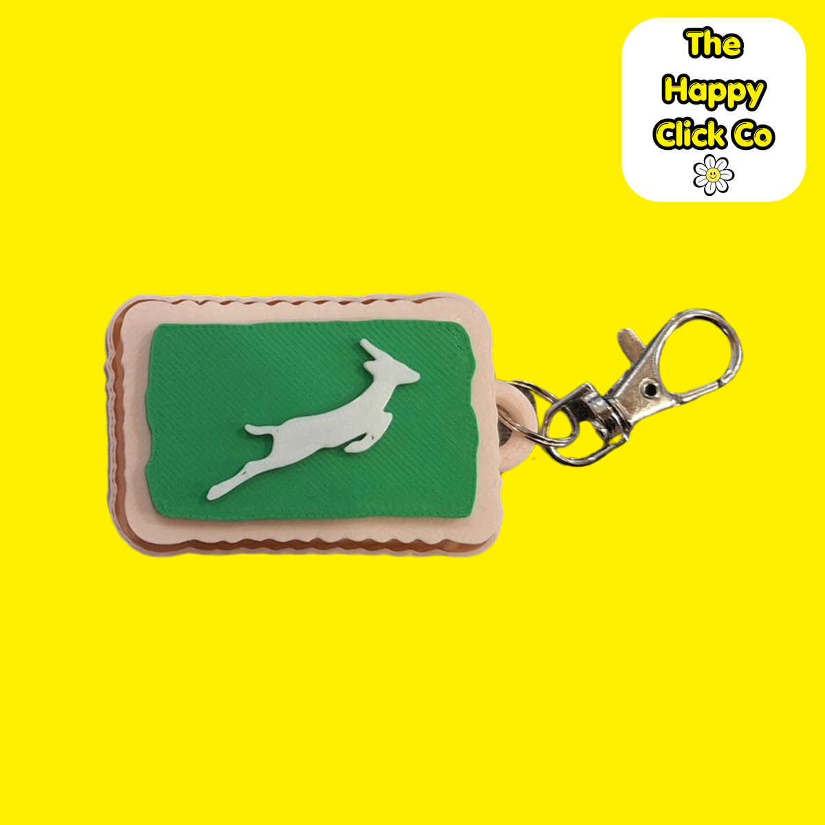 A South African inspired beige zoo biscuit rectangular shaped keychain with a green center panel featuring a white leaping springbok silhouette, attached to a silver metal clasp. The product is photographed on a bright yellow background with a 'The Happy Click Co' logo sticker in the top right corner.