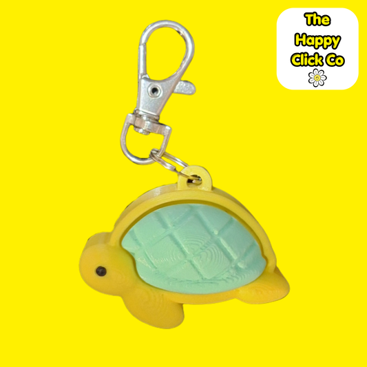 A yellow turtle-shaped fidget clicker keychain with a mint green shell, attached to a silver metal clasp, shown against a bright yellow background with a 'The Happy Click Co' logo in the top right corner.