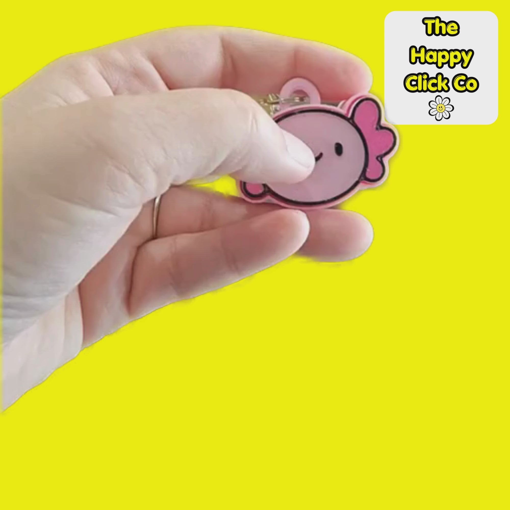 A clicking video of a pink and magenta cartoon axolotl fidget clicker keychain with a silver metal lobster clasp, shown against a solid bright yellow background. A white brand label in the top right corner reads 'The Happy Click Co'.