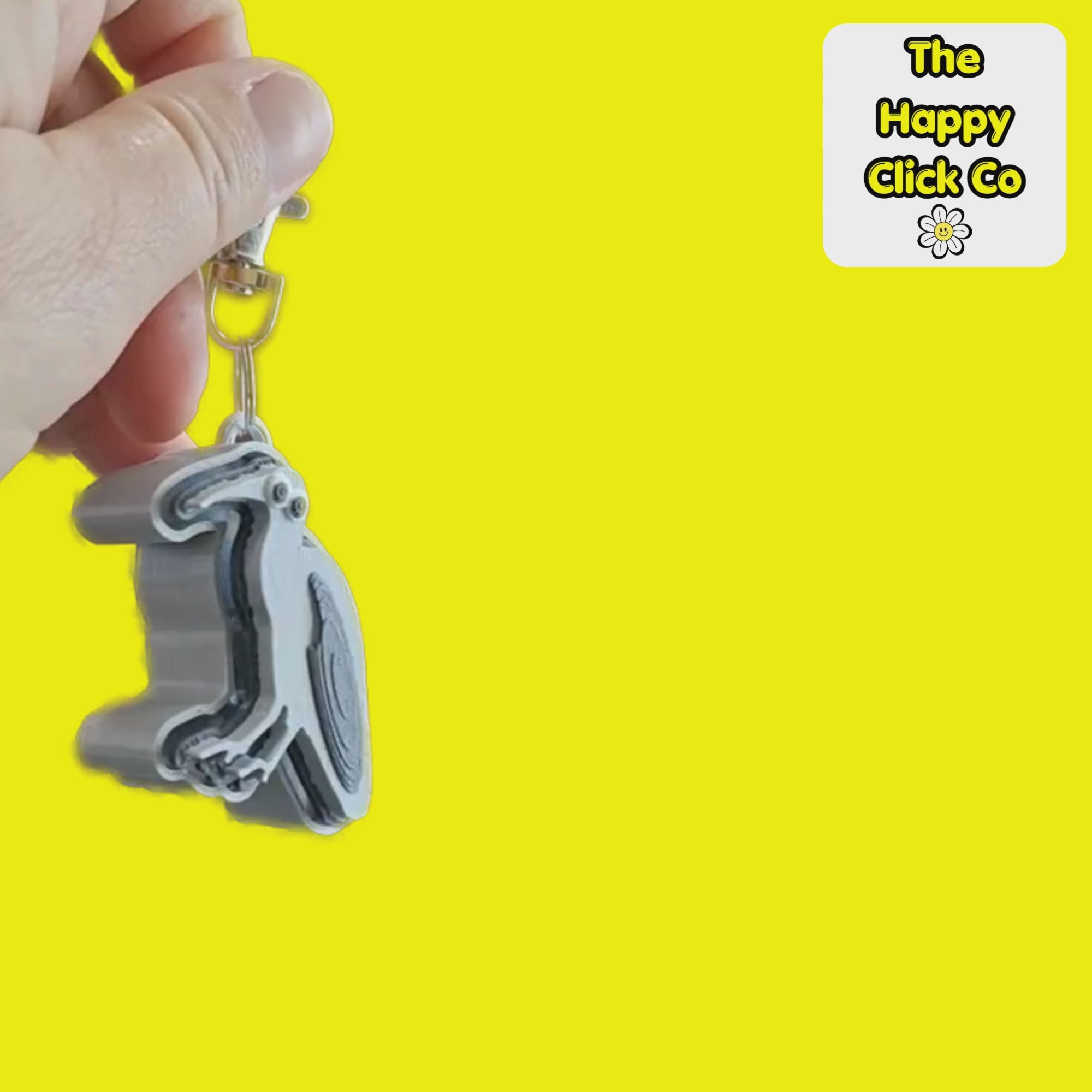 A video of a South African inspired grey and  black Hadeda bird shaped fidget clicker keychain with a silver metal clasp, featuring cartoon eyes on a bright yellow background. A logo for 'The Happy Click Co' is visible in the top right corner. The Fidget Clicker is being dangled and shaken.