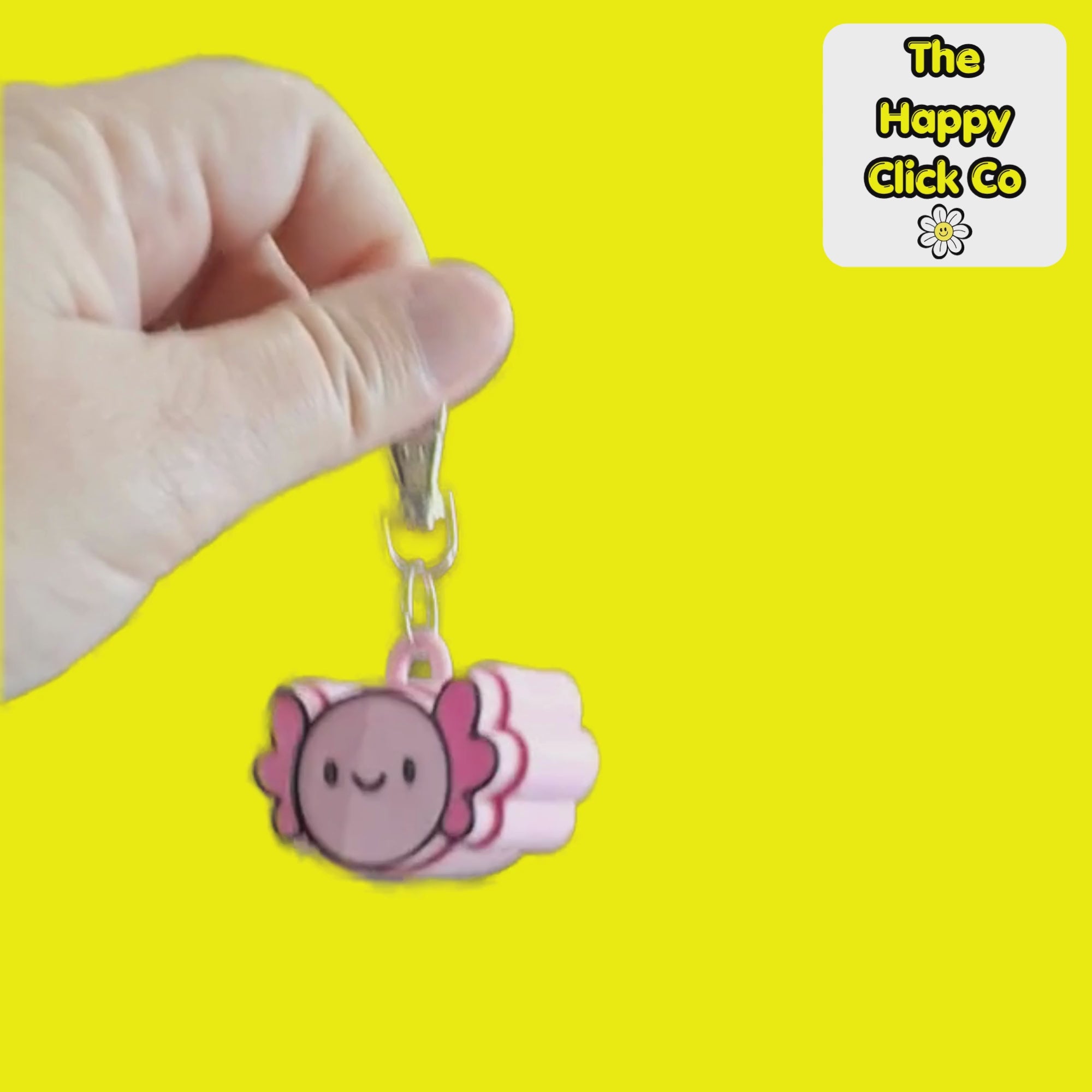 A video of a pink and magenta cartoon axolotl fidget clicker keychain with a silver metal lobster clasp, shown against a solid bright yellow background. A white brand label in the top right corner reads 'The Happy Click Co'. The Fidget Clicker is being dangled and shaken.