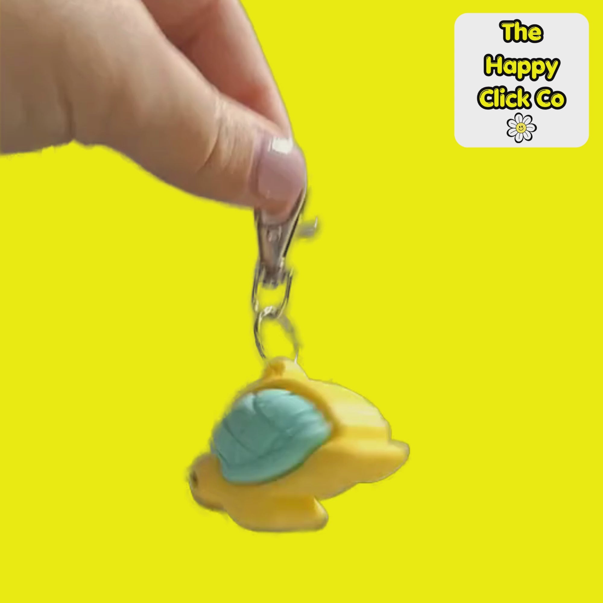 A video of a yellow turtle-shaped fidget clicker keychain with a mint green shell, attached to a silver metal clasp, shown against a bright yellow background with a 'The Happy Click Co' logo in the top right corner. The Fidget Clicker is being dangled and shaken.