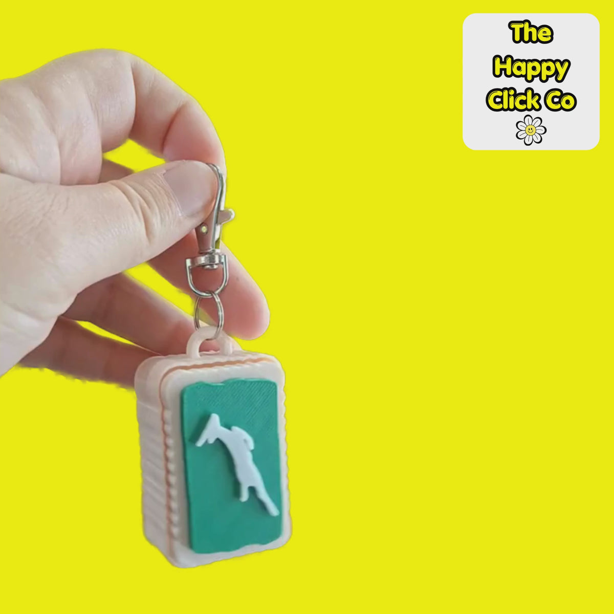 A video of a South African inspired beige zoo biscuit rectangular shaped keychain with a green center panel featuring a white leaping springbok silhouette, attached to a silver metal clasp. The product is photographed on a bright yellow background with a 'The Happy Click Co' logo sticker in the top right corner. The Fidget Clicker is being dangled and shaken.