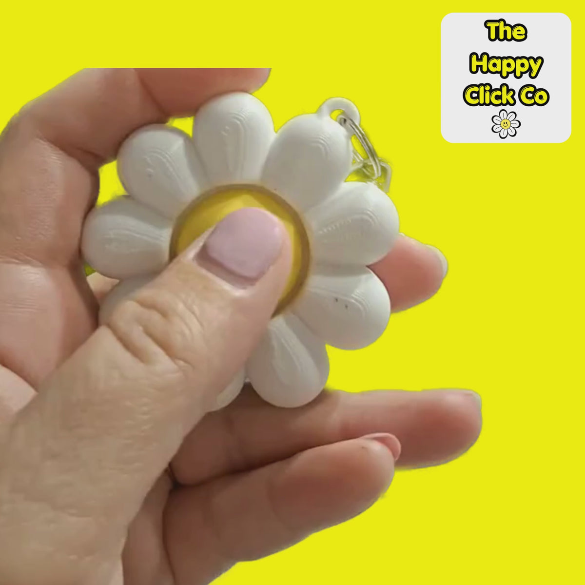 A clicking video of a white daisy shaped  fidget clicker keychain with a smiling yellow face in the center and a silver clasp at the top, shown on a bright yellow background. A 'The Happy Click Co' logo is visible in the top right corner.