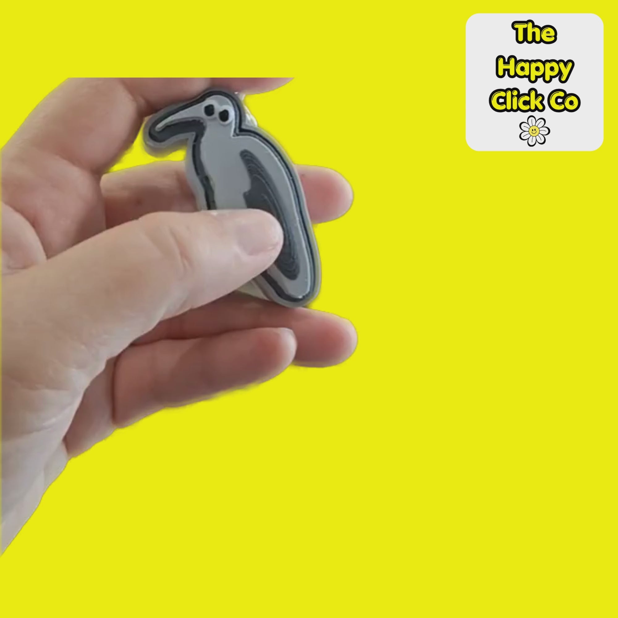 A clicking video of a South African inspired grey and  black Hadeda bird shaped fidget clicker keychain with a silver metal clasp, featuring cartoon eyes on a bright yellow background. A logo for 'The Happy Click Co' is visible in the top right corner.