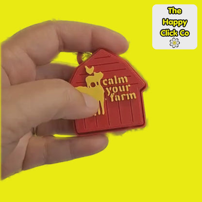 A clicking video of a red barn shaped  fidget clicker keychain with  yellow farm animals and calm your farm text in the center and a silver clasp at the top, shown on a bright yellow background. A 'The Happy Click Co' logo is visible in the top right corner.