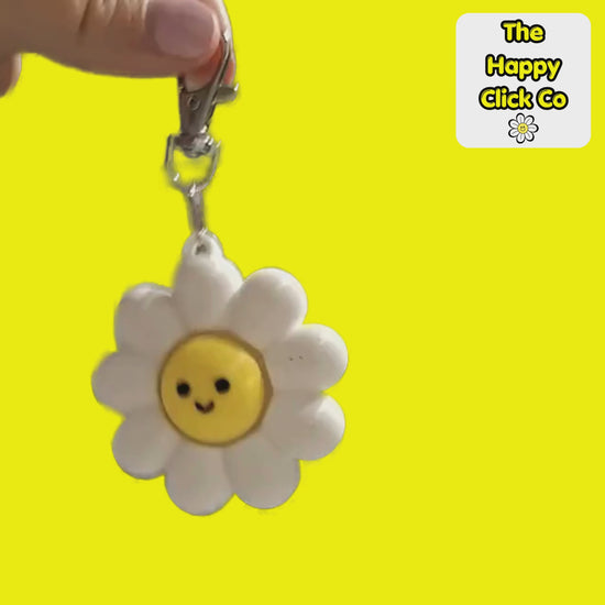 A video of a white daisy shaped  fidget clicker keychain with a smiling yellow face in the center and a silver clasp at the top, shown on a bright yellow background. A 'The Happy Click Co' logo is visible in the top right corner. The Fidget Clicker is being dangled and shaken.