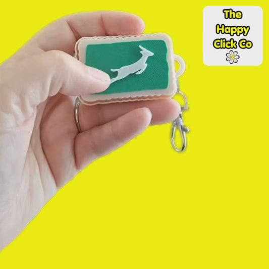 A clicking video of a South African inspired beige zoo biscuit rectangular shaped keychain with a green center panel featuring a white leaping springbok silhouette, attached to a silver metal clasp. The product is photographed on a bright yellow background with a 'The Happy Click Co' logo sticker in the top right corner.