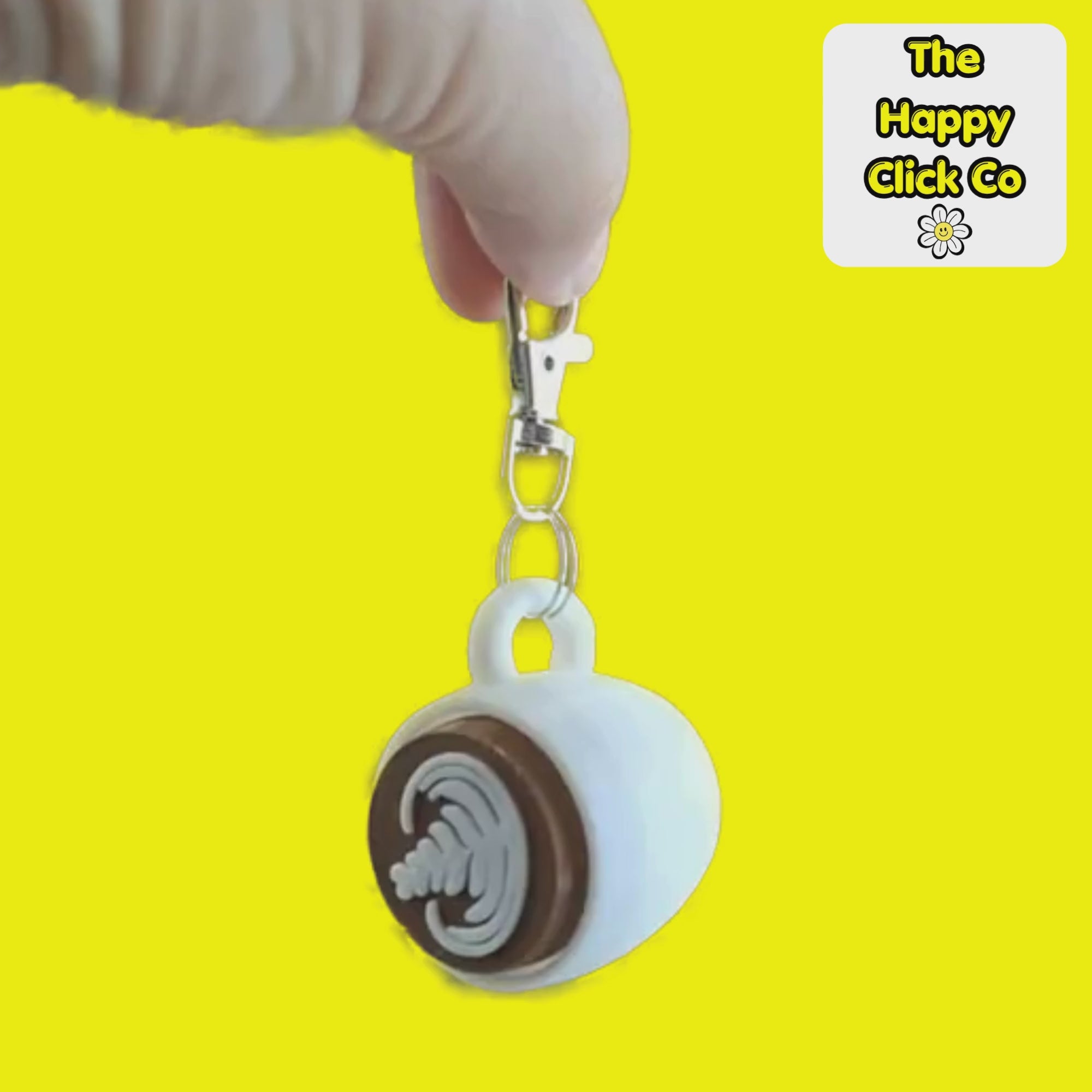 A video of a white coffee cup shaped fidget clicker keychain with a brown centre and cappuccino milk pattern and a silver clasp at the top, shown on a bright yellow background. A 'The Happy Click Co' logo is visible in the top right corner.  The Fidget Clicker is being dangled and shaken.