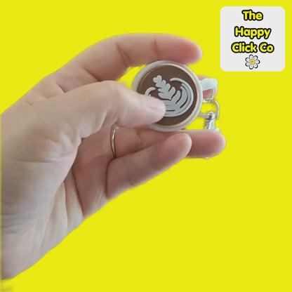 A clicking video of a white coffee cup shaped fidget clicker keychain with a brown centre and cappuccino milk pattern and a silver clasp at the top, shown on a bright yellow background. A 'The Happy Click Co' logo is visible in the top right corner.