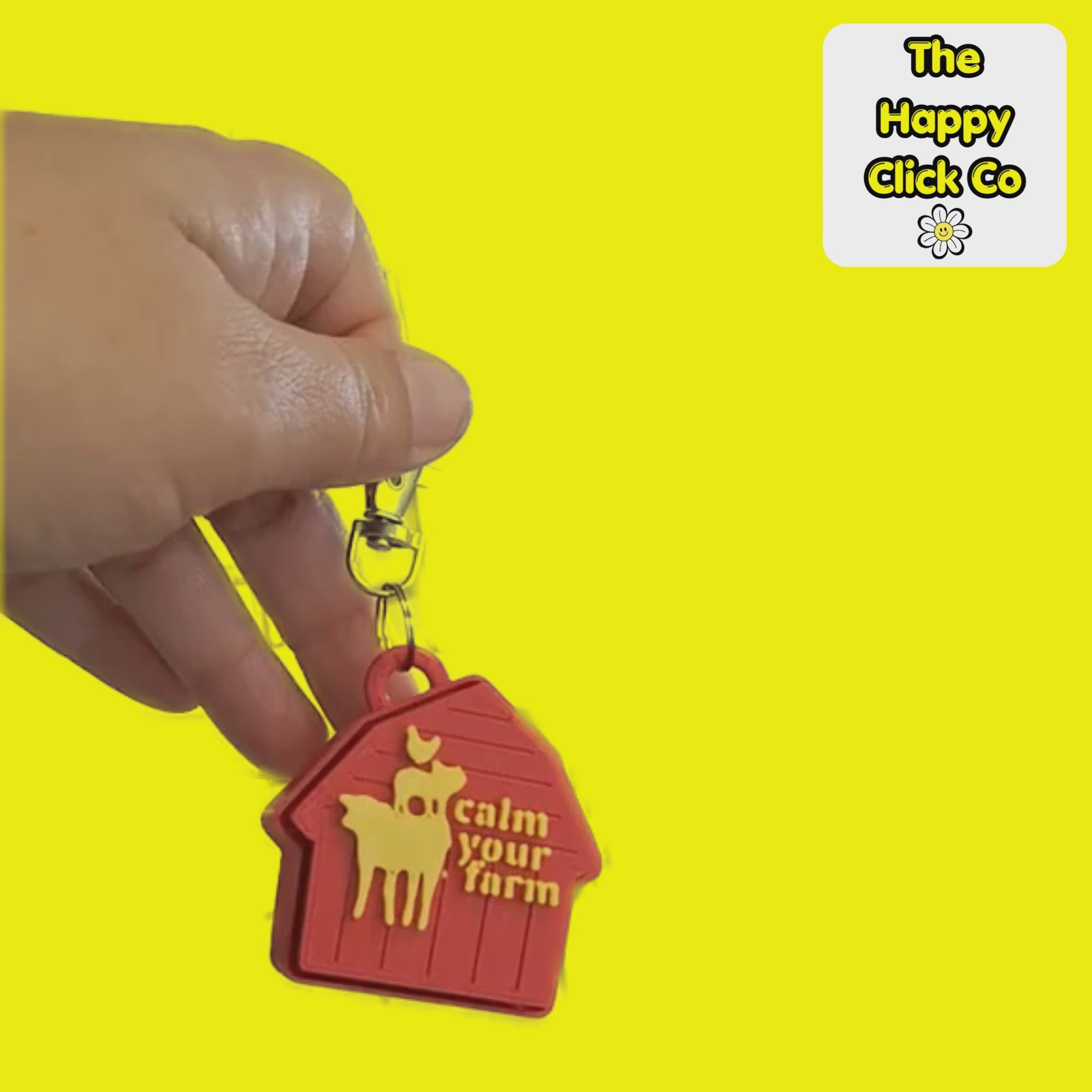 A video of a red barn shaped  fidget clicker keychain with  yellow farm animals and calm your farm text in the center and a silver clasp at the top, shown on a bright yellow background. A 'The Happy Click Co' logo is visible in the top right corner. The Fidget Clicker is being dangled and shaken.